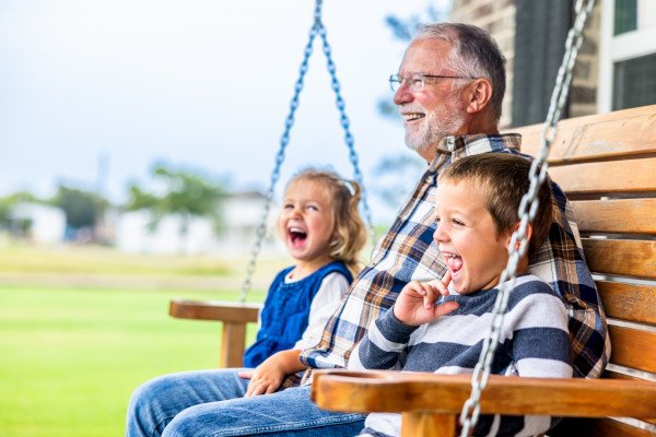 An elderly man and two children sitting on a swing, smiling and enjoying each other's company outdoors.